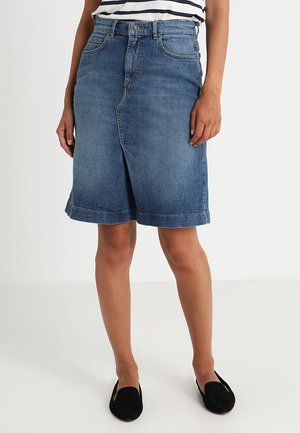 Femme portant une jupe en jean bleue longueur genou avec une fente à l'avant, un haut rayé et des chaussures noires sans lacets, debout sur un sol blanc.