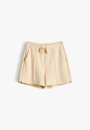 Beige cotton-blend shorts with elastic waistband, drawstring tie, side pockets, and front pleats, laid flat on white background.