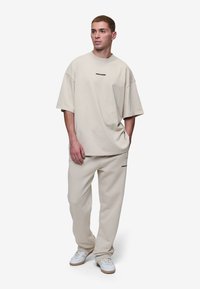 Beige oversize tričko a zodpovedajúce jogger nohavice vyrobené z jemného materiálu, s čiernym logom na hrudi a nohaviciach, spolu s bielymi teniskami.