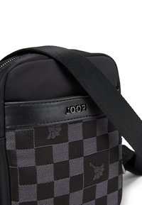 Črno-siva torba za čez ramo z vzorcem šahovnice, gladkim materialom, usnjenimi poudarki in nastavljivim trakom.