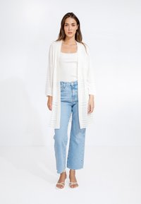 Cardigan bianco a maglia aperta con texture a coste, indossato sopra un top aderente bianco. Jeans a gamba larga blu chiaro e sandali bianchi completano il look.