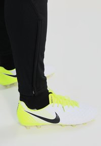 Witte en neon gele Nike voetbalschoenen met zwarte accenten, een gestructureerde bovenkant en een onderscheidend logo; gedragen met zwarte sportbroek.