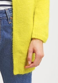 Main reposant à côté de la hanche, portant un pull jaune vif et duveteux et un jean bleu avec une chemise rayée partiellement visible.