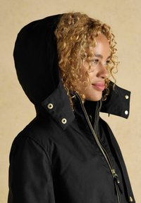 Čierna kapucňová bunda vyrobená z hladkej tkaniny, so zipsom vpredu, zlatými kovovými doplnkami a štruktúrovanou kapucňou s cvokmi.