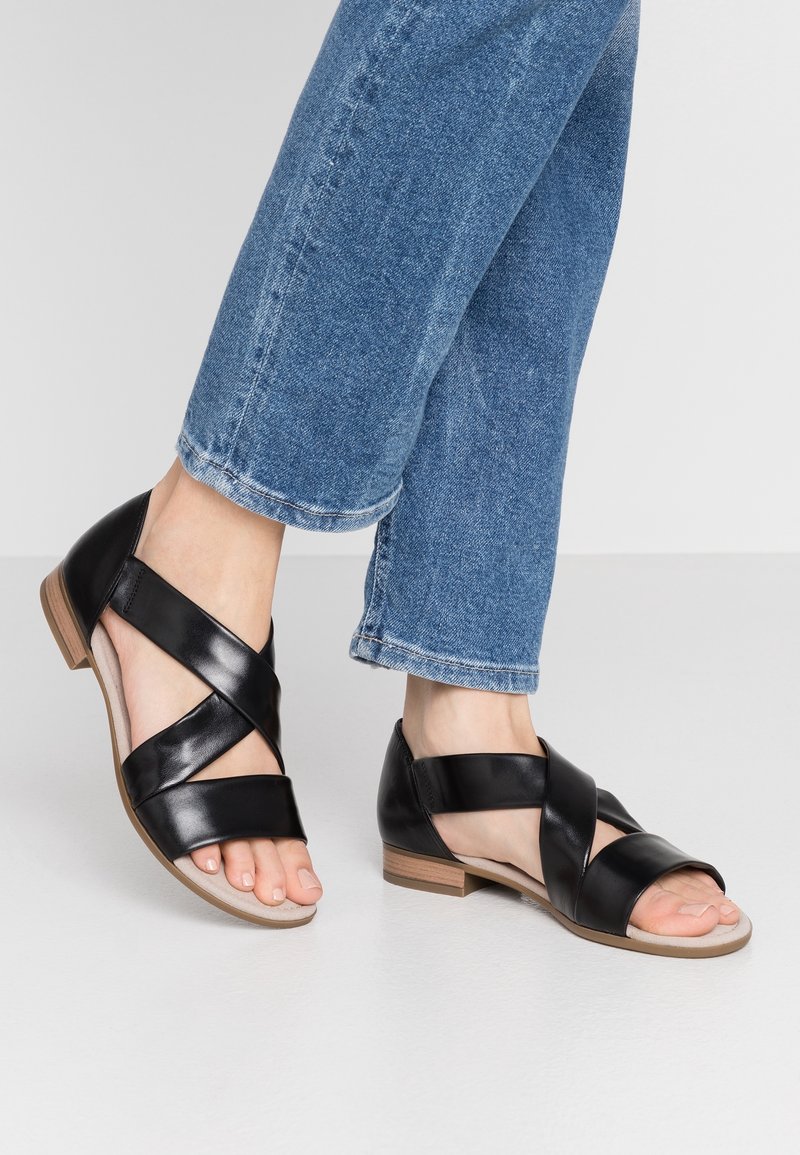 Pieds portant des sandales en cuir noir à brides croisées avec de petits talons en bois, associées à un jean en denim bleu sur fond clair.