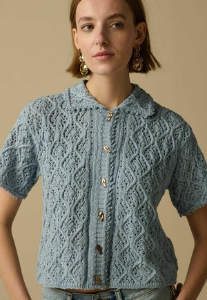 Jeune femme portant un cardigan tricoté à manches courtes bleu clair avec un motif en losange et des boutons dorés, associé à des boucles d'oreilles pendantes dorées.