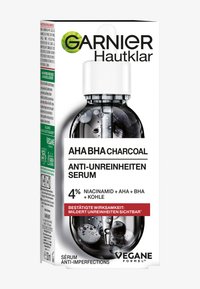 Genomskinlig flaska med svart vätska, vit pipettlock och etiketter på tyska och engelska. Innehåller niacinamid, AHA, BHA och kol.