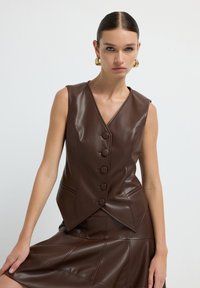 Gilet in finta pelle marrone con scollatura a V, chiusura a cinque bottoni e vestibilità sagomata. La texture liscia e l'orlo asimmetrico aggiungono un tocco moderno.