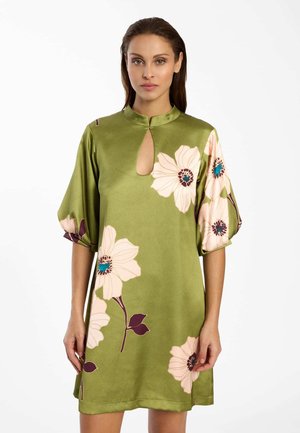 Femme portant une robe en satin vert olive avec de grandes impressions florales rose pâle, col en trou de serrure et manches bouffantes mi-coudes, debout devant un fond blanc.