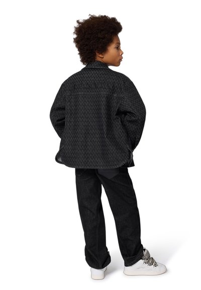 Veste noire à motif en chevron, associée à un pantalon sombre et des baskets blanches avec des lacets noirs et blancs.