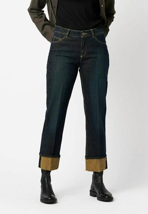 Person wearing dark blue cuffed jeans with brown inner lining, black boots, black top, and olive green jacket, standing against plain background.