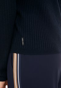 Marineblauer gerippter Strickpullover mit strukturiertem Finish, versehen mit einem kleinen Markenetikett am Saum, kombiniert mit marineblauen Hosen mit vertikalen Streifen.