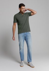 Donkergroene polo met een kraag, gecombineerd met lichtblauwe jeans. Het model draagt grijze sneakers en staat tegen een neutrale achtergrond.
