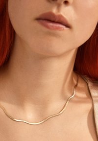 Gold-tone necklace with a wave design; smooth, curved shape; sits close to the neck, highlighting the collarbone area.