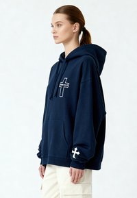 Felpa navy in cotone, con tasca frontale e logo bianco con design a croce. Vestibilità ampia con polsini e orlo a coste.