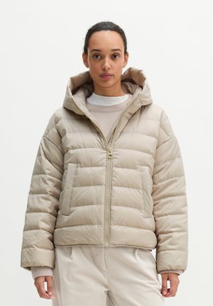 Vrouw draagt een beige pufferjas met capuchon en ritssluiting, gecombineerd met lichtgekleurde corduroy broek en een witte gelaagde trui.