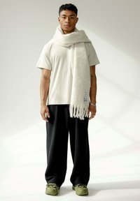 Écharpe en laine blanche avec des franges drapée autour du cou, associée à une chemise légère à manches courtes et un large pantalon noir à côtes. Baskets vertes.