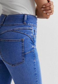 Niebieskie dżinsy z wysokim stanem, kontrastującymi pomarańczowymi szwami i dopasowanym krojem, z widocznymi kieszeniami i delikatną fakturą.