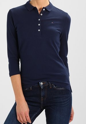 Vrouw draagt een marineblauw poloshirt met lange mouwen en knopen, gecombineerd met donkerblauwe spijkerbroek, tegen een effen achtergrond.