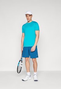 A turquoise athletic t-shirt with a round neck and short sleeves, paired with navy shorts. A white cap and tennis racquet complete the look.