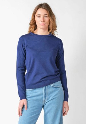 Jeune femme aux cheveux châtain clair portant un pull bleu à manches longues et un jean bleu clair, debout devant un fond blanc uni.