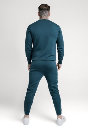 Mann mit Hals-Tattoo trägt dunkelgrünen Jogginganzug und weiße Turnschuhe, steht mit dem Rücken zur Kamera vor schlichtem hellem Hintergrund.