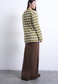 Femme debout contre un mur blanc, portant un haut à manches longues rayé olive et blanc et une longue jupe marron, regardant sur le côté.