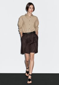 Kvinde går iført en beige knap-åben skjorte, højtsiddende mørkebrun shorts med et sort flettet bælte og sorte ankelrem-sandaler.