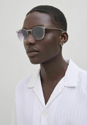 Gafas de sol redondas con montura transparente y lentes oscuros degradados; combinadas con una camisa blanca a rayas que presenta un cuello y detalles de botones.