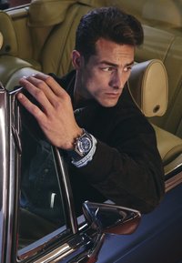 Orologio analogico con faccia blu e accenti argentati su un cinturino scuro, abbinato a una giacca di jeans e a una giacca in suede marrone. All'interno di un'auto con sedili in pelle beige.