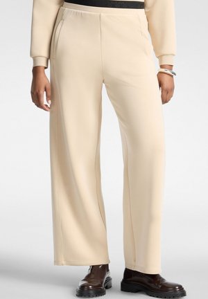 Pantaloni beige a gamba larga indossati con stivali neri in pelle e una felpa beige, con tasche frontali e vita visibile.