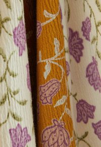 Due pezzi di tessuto: uno crema con motivi floreali verdi e viola, l'altro arancione con accenti floreali viola. Superfici tessurate e decorate.