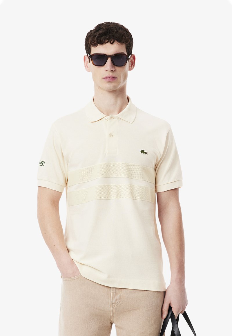 Polo en crème avec un design de rayures horizontales texturées. Dispose d'un logo vert sur la poitrine et d'un col classique.