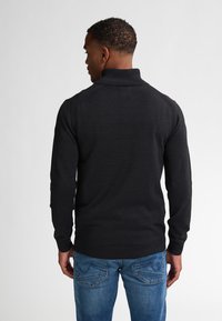 Schwarzer Strickpullover mit hohem Kragen, gerippten Akzenten an den Schultern und langen Ärmeln, getragen über blauen Jeans. Glatte Textur.