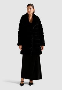Abrigo de piel sintética negro con solapas anchas, textura acanalada y un ajuste holgado, superpuesto sobre un vestido largo negro y con zapatos de punta.