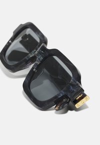 Überdimensionierte Sonnenbrille mit einem dunkelgrau marmorierten Rahmen, großen getönten Gläsern und goldenen Beschlägen mit den Buchstaben "RSF" an der Seite.