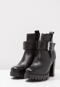 Botas de cuero negro hasta el tobillo con un tacón grueso, que cuentan con una hebilla lateral y suelas texturizadas, exhibidas sobre un fondo blanco liso.