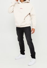 Felpa beige con tasca frontale e piccolo logo rosso, abbinata a jeans neri slim e sneakers bianche. Design semplice, informale e moderno.