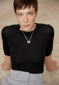 Black sheer short-sleeve top worn with a silver heart pendant necklace. Light gray high-waisted pants with a textured fabric.