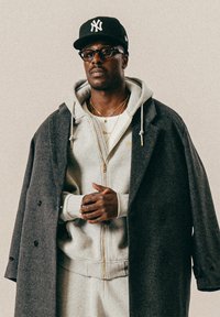 Homme portant une casquette des Yankees noire, des lunettes, un hoodie gris zippé sous un manteau gris foncé, avec des colliers en or et les mains jointes.