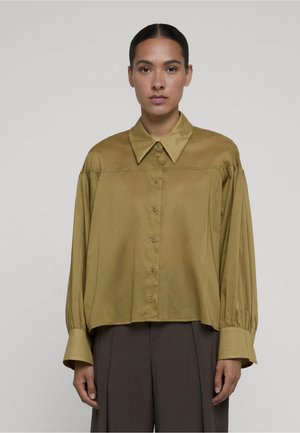 Mujer con cabello oscuro en un moño bajo, vistiendo una camisa de botones amarilla mostaza suelta con mangas largas y pantalones marrón oscuro, de pie frente a un fondo liso.