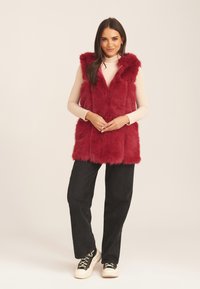Red faux fur vest with a hood, featuring a v-neck design, layered texture, and worn over a light pink long-sleeve top and black pants.