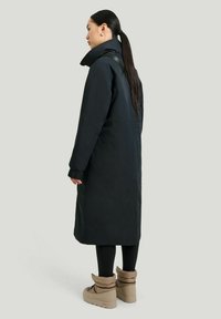 Lång svart kappa med hög krage och elastiska manschetter, som matchas med ljusbruna stövlar med en mjuk, vadderad textur.