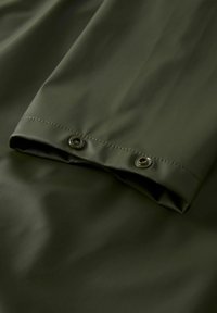 Sötétzöld vízálló anyag sima textúrával, hajtott mandzsettával, amelynek alján két fém patent található.