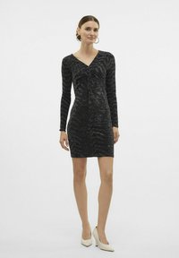Vestido negro de manga larga con escote en V y detalle de fruncido en el frente, que presenta un patrón texturizado. Combinado con tacones puntiagudos de colores claros.