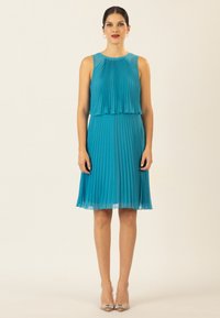Ein ärmelloses, türkisfarbenes, plissiertes Kleid mit gestaffeltem Design, fließendem Saum und seitlichen Taschen. Kombiniert mit klaren, verzierten High Heels.