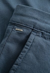 Mėlynos spalvos raštuota kelnėms skirta medžiaga su tekstūruotu audimu, juosmens juostoje esanti sidabrinė metalinė etiketė su užrašu „GABBA“.