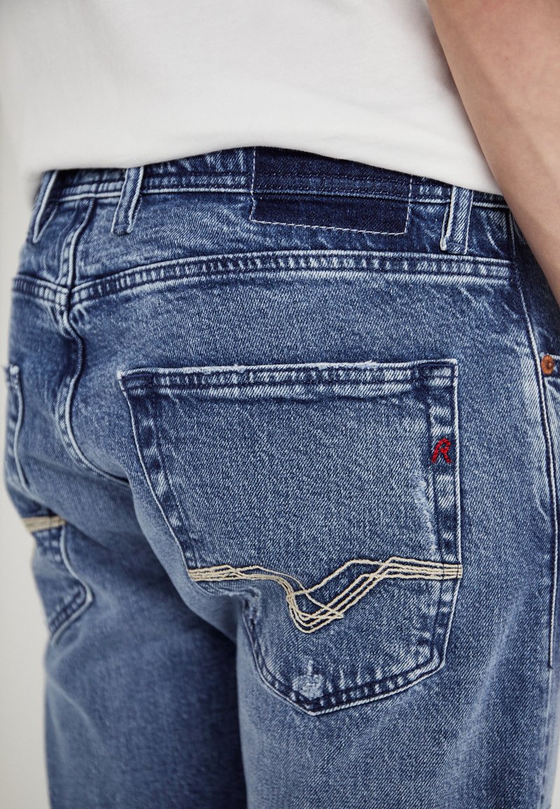Blauwe denim jeans met een vervaagde afwerking, achterzakken met crème stiksel en een klein rood logo op de rechterzak.