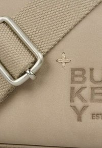 Beige kanvas taske med en sølvmetalknap og præget brandnavn, med detaljeret syning og en tekstureret overflade.