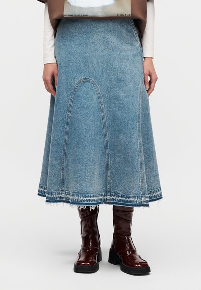 Φούστα από ανοιχτό μπλε τζιν με φαρδιά γραμμή και ακατέργαστο τελείωμα. Συνδυάζεται με λαμπερά μπορντό μποτάκια αστραγάλου, εμφανιζόμενη από τη μέση και κάτω.